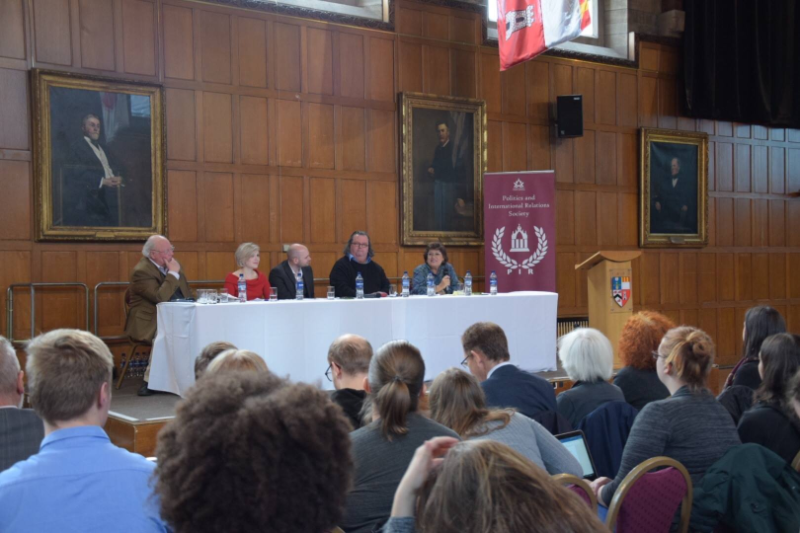 A panel of speakers sit in a wood panelled room in front of an audience