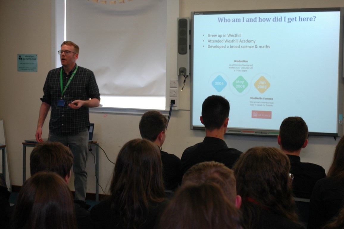 Dr. Alan McCue is giving a presentation to school pupils. The screen he is reading from reads "Who am I and how did I get here?"