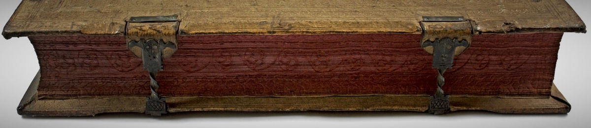 The edge of a thick book held together by two metal clasps. The edging is decorated with spiral and floral details.