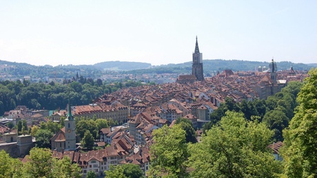 a city with red tiled roofs on lots of buildings, a dome in the background and multiple towers with spires. There are blue sky and tree covered hills behind the city and in the foreground there are trees.