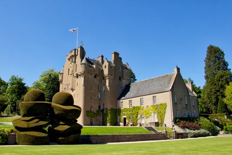 A side on view of Crathes Castle. It is a very sunny day with clear blue skies. There are lots of plants and trees in the foreground.