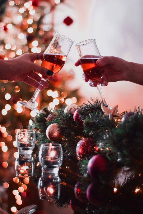 Two people clink glasses of a red cocktails in champagne glasses. Behind them are festive decorations, such as a fir garland and fairy lights.