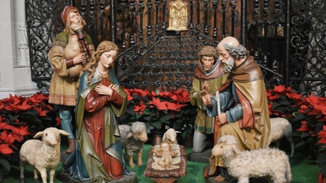 figurines from the nativity, in a traditional style, arranged in a scene in front of black metal gates and surrounded by red poinsettias.