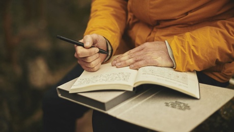 a close up of a persons hands on top of two open notebooks with a pen in one hand ready to wrtie. the person seems to be sitting outdoors and is wearing a yellow rain coat.