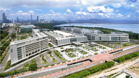 a view from above a complex of modern white buildings, surrounded by grenery and roads, with a city in the background and mountains behind the city, a blue cloudy sky above.