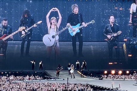 Taylor Swift, a tall woman in a metallic dress and silver guitar, poses with her hands in a heart on stage. She is joined on stage by three guitarists and a bassist, all in dark clothing.