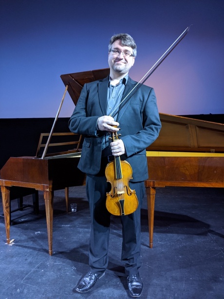 Dr Aaron McGregor in a blue shirt and suit, holding a stringed instrument, in front of a piano.