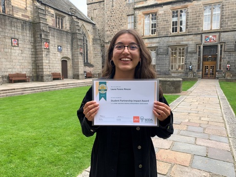 A smiling Laura holding up her certificate against the backdrop of King's Chapel