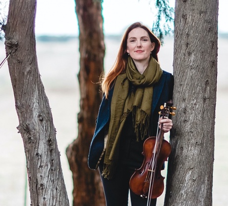 A red haired woman in a green scarf and blue blazer is stood against a tree whilst holding a violin. She is smiling.