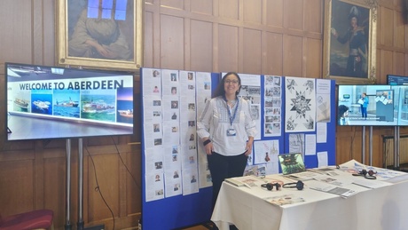 Dr Alejandra Rodriguez-Remedi presenting at the Elphinstone Hall, with poster boards behind and a table with infomration in front.