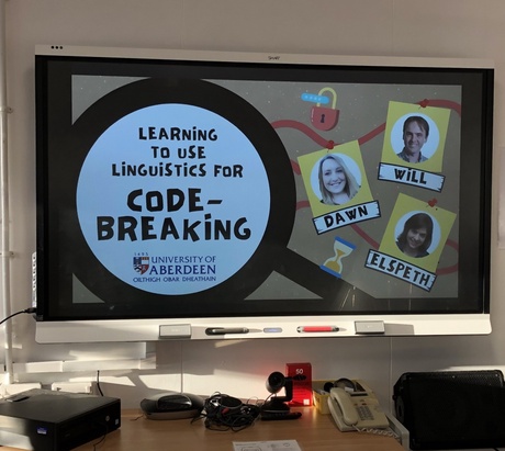 A projector screen, with the details of Linguistics lecturers and information about the code breaking course on it.