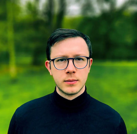 James Aburn - A white man in in a dark jumper, dark hair and circular glasses in a green grassy field.