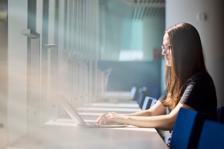 A female student is seated in a lit corridor, typing on a laptop.