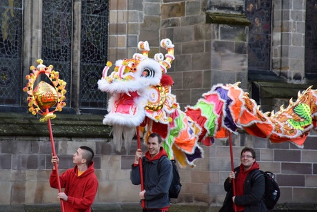 A photograph of 2 people holding a Chinese Dragon above them, behind another person outside