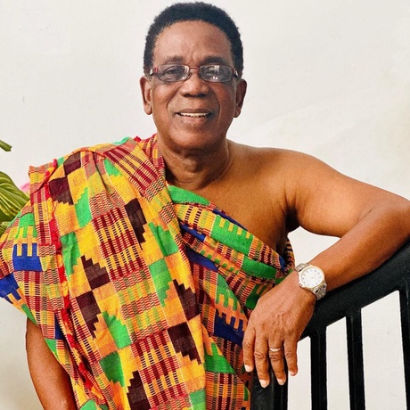 A Ghanaian man with glasses in smiles for the camera wearing a kente cloth outfit