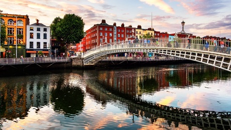 A river through a city, with a white metal bridge, lined with colourful houses and pedestrians.