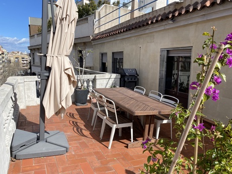 A sunny rooftop terrace with a large dining table, a barbeque, a folded parasol, and some purple flowers.