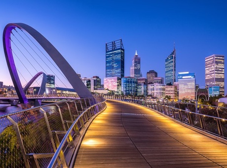 A photo of the Perth skyline taken from a long wooden boardwalk. It is dusk and the sky is a dark blue and the lights of the city are shining in contrast.