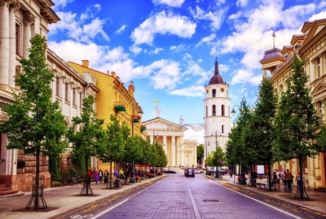 image of a paved city street. the street is lined with tree's, lamp posts,and old stone buildings. at the end of the street is a whtie stone church with columns and a tall octagonal bell tower.