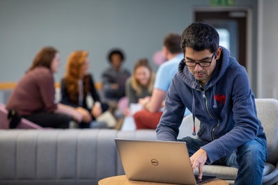 Individual sitting on a sofa and using a laptop. There is a small groip of people meeting in the background.