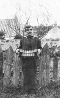 Old black and white photo of young mustachioed man playing concertina in front of a fence.