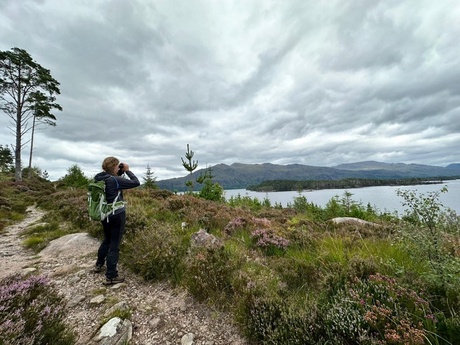 women with binoculars looking across a heather landscapes with trees and a cloudy sky