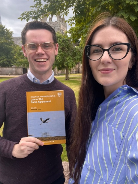 A photo of Dr Daria Shapovalova and Dr Mitchell Lennan on King's College Campus, Dr Lennan is holding the Research Handbook on Law of the Paris Agreement