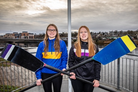 Two people standing in front of the River Dee, each holding an oar