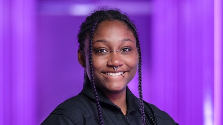 Anastatia smiling at the camera wearing a black shirt in front of a purple background.