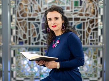 Professor Alexandra Johnstone in a blue dress with a book in her hand in front of a stained glass window