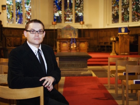 Professor Mealor in a black suit and tie in front of stained glass windows in a chapel