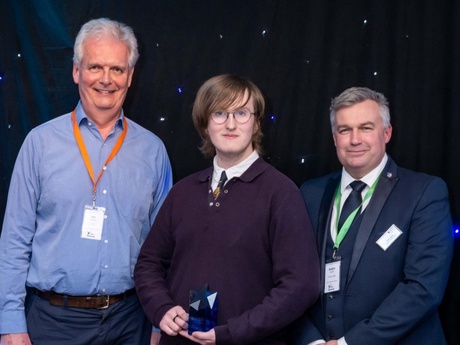 One of the winning Mackie Academy pupils holding their award and flanked by YE Grampian chairman Ian Phillips and Adam Smith from the University Business School, set against a dark background with glitzy lights