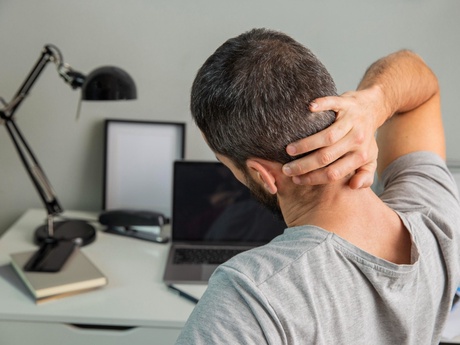 A man at a desk with a laptop leaning back rubbing his neck