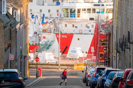 Image of a woman walking across a street in Aberdeen with ships in the harbour in the background