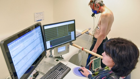 A patient volunteer wearing an oxygen mask walks on a treadmill while a researcher monitors data on a monitor