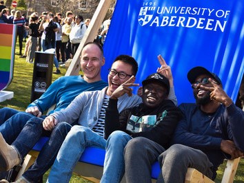 A group of four smiling students sharing a deck chair on the lawn at Old Aberdeen campus