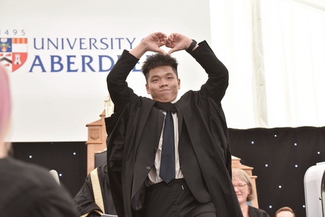 Man on graduation stage making a heart with his hands