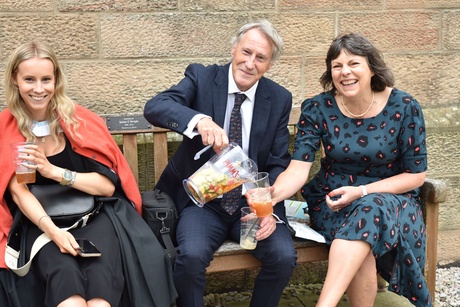 Three people on a bench with a man in the middle holding a drinks pitcher