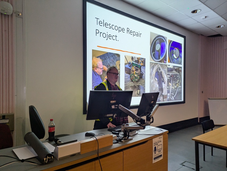 A presenter giving a talk in the Zoology Lecture Theatre. On the screen is a slide about a project to repair the University's 11" telescope.