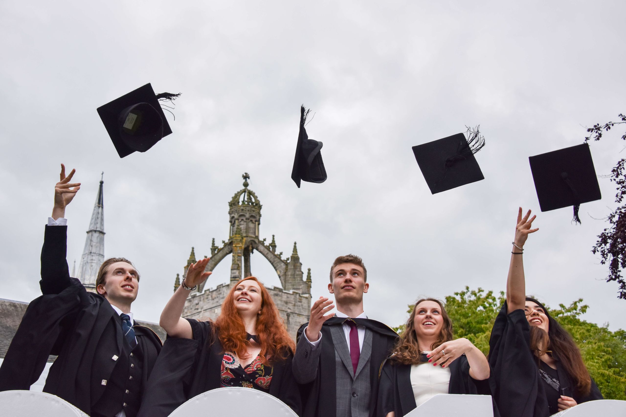 Five students celebrating graduation day at University of Aberdeen by throwing their graduation caps in the air..